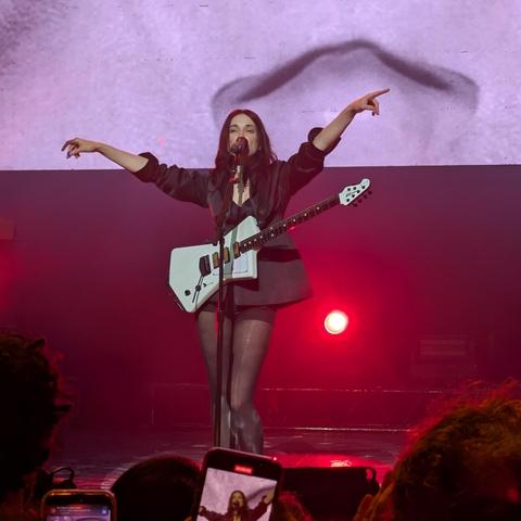 Una mujer de cabello oscuro y largo, con un vestido negro corto y chaqueta negra, está de pie en un escenario iluminado con luces rojas. Está tocando una guitarra eléctrica blanca y cantando a un micrófono. Sus brazos están extendidos, uno apuntando hacia arriba y el otro hacia el lado, mostrando una pose dinámica y enérgica. El fondo es una pantalla grande con un tono púrpura claro y una sombra difusa. Se ven partes de la audiencia, con teléfonos móviles grabando la actuación. La atmósfera general es la de un concierto con mucha energía.