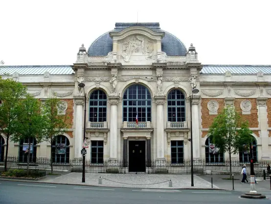 The image depicts an ornate building, characterized by its grand facade, intricate architectural details, and large arched windows. It features decorative elements above the entrance and a diverse texture with both stone and brick materials. The structure is likely situated in an urban setting, surrounded by trees and street elements.

Image Credits: Wikimedia / PHGCOM / Public domain