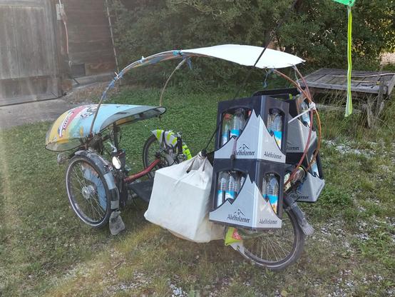 Das Liegedreirad mit Wind- und Regenschutz und Dach beladen mit vier leeren Wasserkästen und noch seitlich einer Tasche mit Leergut