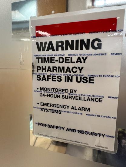 A “Warning: time-delay pharmacy safes in use” sign, with a sheet overlaying the words with the words “Remove to expose adhesive”. The sign is held against a frosted glass window by two broad transparent tape, which has made the frosted glass clear behind them.