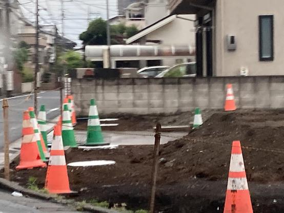 A street scene featuring several traffic cones in orange and green, surrounding a construction area. There is a dirt patch in the foreground, with a wall and buildings visible in the background.