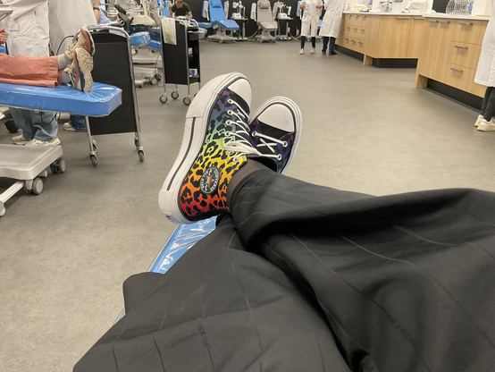 Emma zit ontspannen met haar benen gekruist tijdens het doneren van plasma bij de bloedbank. Ze draagt Converse All Stars met een regenboog panterprint.