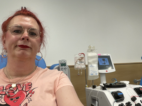 Emma, met rood haar en bril, glimlacht ontspannen tijdens haar plasmadonatie bij de bloedbank. Achter haar zijn medische apparaten te zien die gebruikt worden voor het afnemen van plasma. Ze draagt een roze t-shirt met een hart en een Nike-logo en de tekst "Love is in the AIR"