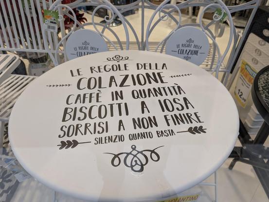 A decorative round white table is displayed in a store, featuring playful black typography with the following Italian text:

"LE REGOLE DELLA COLAZIONE
CAFFÈ IN QUANTITÀ
BISCOTTI A IOSA
SORRISI A NON FINIRE
SILENZIO QUANTO BASTA"

Translation:
"The rules of breakfast
Plenty of coffee
Lots of cookies
Endless smiles
Silence, just enough"

The table is part of a matching white wrought iron bistro set, including chairs with circular backrests that also feature the phrase "LE REGOLE DELLA COLAZIONE" - The rules of breakfast. The setting is inside a home decor or furniture store, with price tags and other products visible in the background.