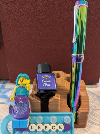 Shiny rainbow hued Conklin fountain pen with Cosmic Glow ink bottle on stand. In the foreground a LEGO mermaid sits on a Leece name badge.