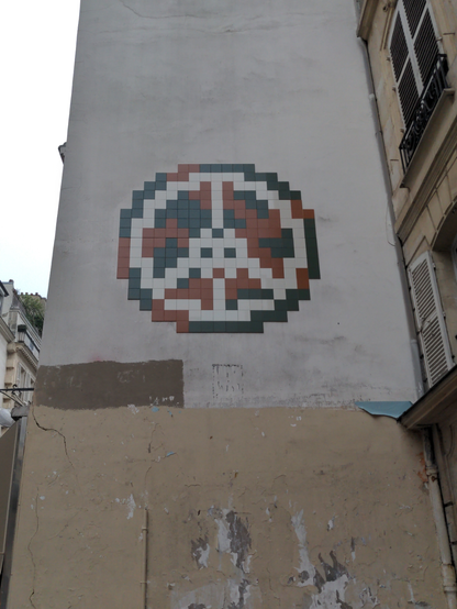 Sur un haut pignon de mur, un grand invader au milieu du symbole de la paix, tracé en blanc sur fond rouge et noir, en mosaïque dans le style d'Invader.