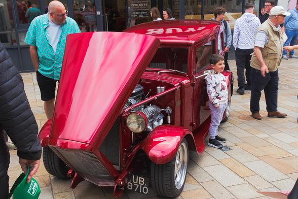 The image captures a vibrant street scene centered on a shiny red custom hot rod with its hood dramatically raised, revealing a polished engine. A young boy stands beside the car, smiling proudly with his hands in his pockets. He's dressed in a light grey outfit with playful prints. People of various ages gather around, admiring the car. One man in a turquoise shirt leans in for a closer look. Behind them, a “Five Guys” restaurant and its glass storefront provide an urban backdrop. The mood is cheerful and lively, filled with curiosity and shared appreciation for the unique vehicle on display.
