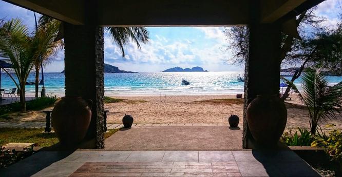 A view from a shaded porch looks out onto a sunny beach with turquoise water. Two large, dark, round pots flank the entrance. Palm trees and other greenery are visible on either side. In the distance, small islands dot the horizon under a partly cloudy blue sky.
