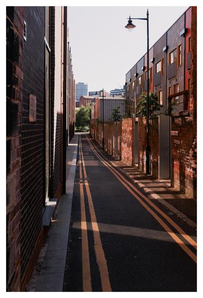A narrow alleyway flanked by tall buildings stretches into the distance. The left side is a dark brick wall with a street sign reading “Cotton Street.” The right side has red-toned brick walls and newer-looking residential windows. A streetlamp stands tall on the right. Twin yellow lines run down the middle of the dark tarmac, pulling the eye toward a patch of green trees at the far end. Soft sunlight casts long, dramatic shadows across the scene. The alley is empty, quiet, and bathed in the warm glow of either morning or late afternoon light. The mood is calm and thoughtful