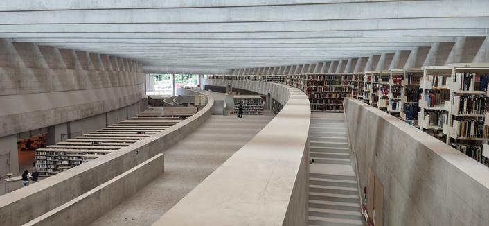 Photo prise depuis une gallerie à l'intérieur du bâtiment. Il est vaste et lumineux. Le béton est omniprésent. On voit l'alignement de nombreuses étagères à livres