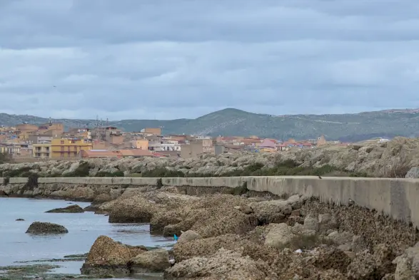 bild im querformat im hafen.
rechts im bild die kaimauer, dahinter die stadt licata und eine bergkette mit wolken darüber.
im rechten bilddrittel seiht man ganz klein einen eisvogel auf der kaimauer sitzen