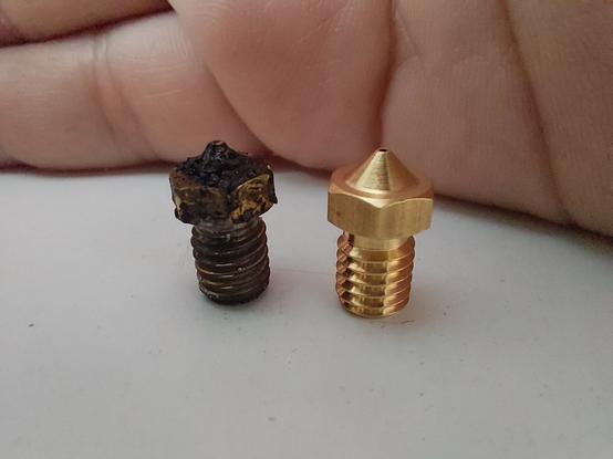 Two 3D printer nozzles on a white table with my beautiful hand in the back blocking out the background for the photo. The left brass nozzle is very dark and has a "patina" of filament shmoo on it. The right one is in perfect condition, shining in a bright gold-like brass color.