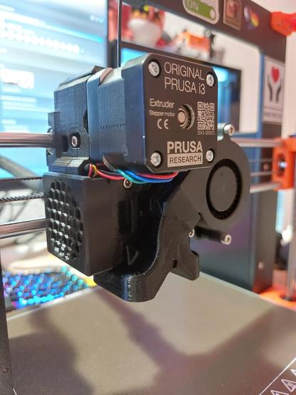Modified Prusa Mk3S+ tool head. In the background you can see a blurred computer screen. Tye tool head has its filament fan mounted vertical on the right side instead of tilted forward in the center. The fan duct is custom printed from black ASA and looks like an inverted Y-shape. The "prongs" on the bottom part surround the nozzle that is hidden behind it.