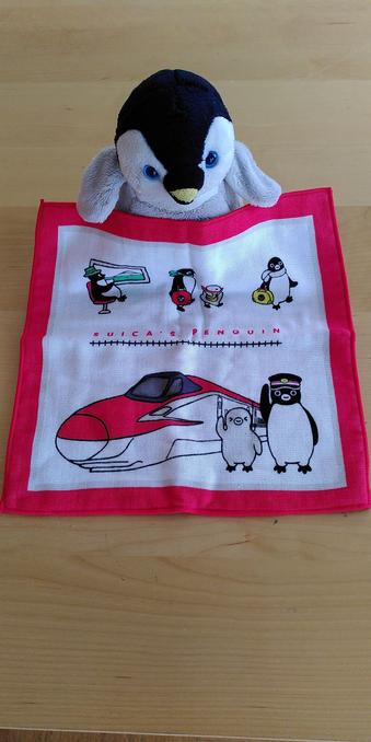 Archy sitzt am Tisch vor ihm liegt ein kleines Handtuch das er mit den Flossen hält. Das Handtuch ist ca 20x20 Zentimeter groß und ist weiß mit rotem Rand. Am Handtuch ist ein Shinkansen abgebildet neben dem zwei Punguine stehen. Über dem Zug steht "Suica's penguin" darüber sind reisende Pinguine zu sehen.