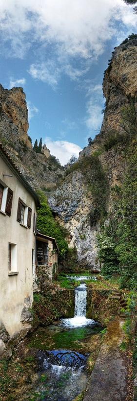 A vertical, wide-angle shot captures a picturesque scene in what appears to be Moustiers-Sainte-Marie. On the left, the side of a light-coloured building with brown-shuttered windows is visible. To the right, a stream flows gently, cascading over small weirs to form tiny waterfalls. The water is clear, revealing the stones beneath. Lush green vegetation lines the banks of the stream.
Towering above are craggy, light-coloured cliffs, with patches of greenery clinging to their sides. In the distance, nestled higher up on the rock face, a small building with a steeple can be seen against a backdrop of a partly cloudy blue sky. The overall impression is of a charming, natural landscape integrated with human habitation.