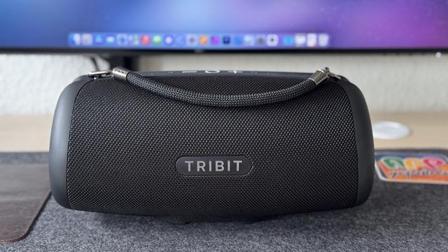 Tribit StormBox Lava mitsamt Trageschlaufe steht auf einer dunklen Filz-Schreibtischunterlage vor einem Monitor, auf dem unscharf ein Mac-Dock zu sehen ist. Am rechten Bildrand ist ein appgefahren-Bierdeckel zu sehen.