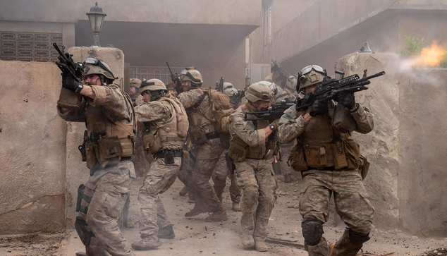 Een aantal Amerikaanse soldaten komen uit de poort bij een Irakese woning, ze liggen onder vuur en schieten zelf ook