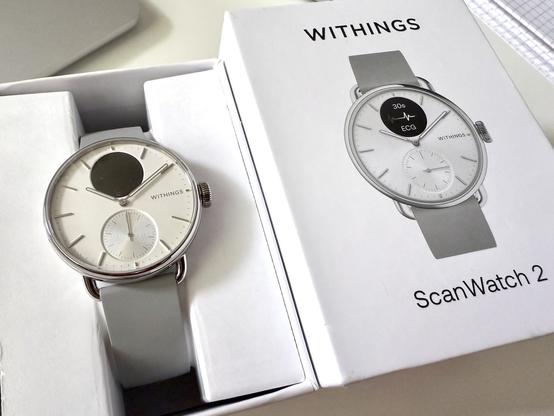 Auf dem Foto ist die Withings ScanWatch 2 in der 38mm Version zu sehen. Uhr in Verpackung neben dem Deckel. Zifferblatt der Uhr ist gelblich im Gegensatz dazu die Abbildung auf der Verpackung hat ein weißes Zifferblatt.