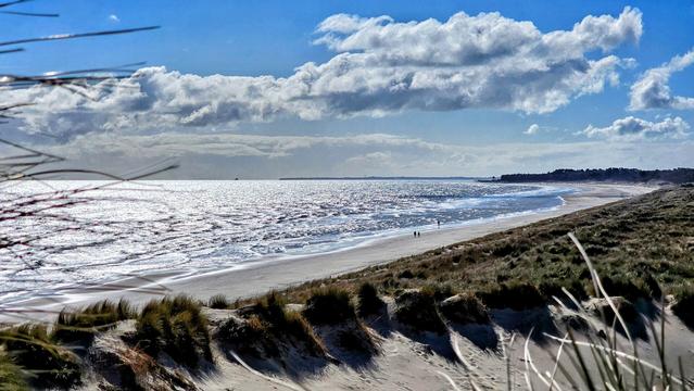 A wide view of an expansive beach on the East Coast of Ireland, featuring bright, sparkling water, a wide sandy shore, and grassy dunes in the foreground. Two small figures are walking along the water's edge, with a distant landmass visible. The sky is partly cloudy, and the sunlight creates reflections on the sea.