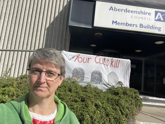 Maggie standing outside the Aberdeenshire Council building. There’s a banner that says “your cuts kill” on the railings behind her