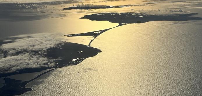 A sunrise aerial view of the islands Miquelon-Langlade, with the smaller Ile-St-Pierre in the background, with light low cloud cover. The ocean colours are orange-yellow in the center third of the photo, fading to steel grey nearer the right and left edges. The islands are dark grey, but ponds and inlets take on the brighter ocean colours. The large island spans from the bottom left corner to the top right corner, with only a small neck of land connecting the two larger portions. [over the northwest Atlantic, December 2024]