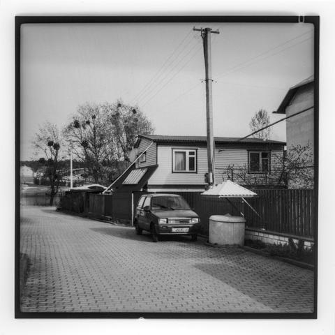 This is a black-and-white photograph of a quiet residential street. The road is paved with interlocking bricks and runs alongside a wooden fence. A small car is parked near the fence, next to a covered well. Behind the car is a two-story house with siding, multiple windows, and a satellite dish mounted on the side. There are utility poles with wires stretching across the scene. Leafless trees, some with large clumps of mistletoe, are visible in the background, indicating an early spring setting. The overall mood of the photo is calm and slightly nostalgic, enhanced by the monochrome tone and film-like aesthetic.