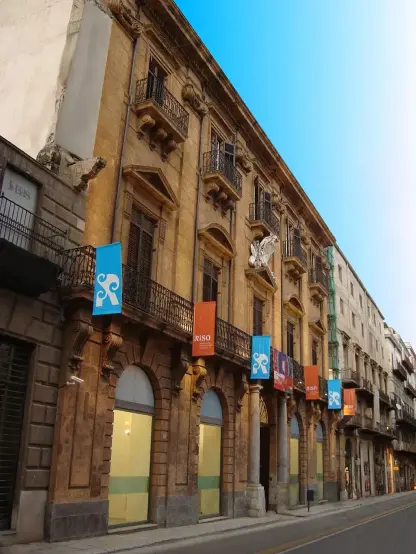 The image shows a building with an elaborate architectural design. It features balconies with wrought iron railings and decorative elements on the facade. There are several banners hanging from the building, some of which have a logo or symbol, and the street appears to be quiet with no visible people. The sky above is blue, suggesting a clear day.

Image Credits: Wikimedia / PacoSoares di Wikipedia in italiano / Public domain
