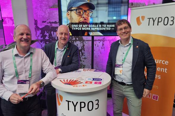Three men stand at a TYPO3 booth with branded materials, smiling for the camera. A screen and TYPO3 banners are visible in the background. Photo: Mike Gifford