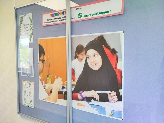 A school bulletin board displays posters and photographs. One photo shows a student in a black hijab and purple sweater working at a desk with papers and colored pencils. Another photo shows a student in glasses holding a utensil at a table. The board also features motivational signs with messages about respect, sharing, and support, as well as a small illustrated sign with positive affirmations.