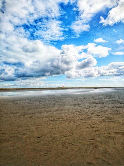 Weites Watt an der Nordsee, mit ebenso weitem blauen Himmel mit großen weißen Wolken. Am Horizont ein Leuchtturm, winzig klein.