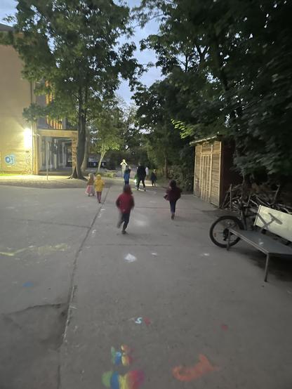 Das Bild zeigt verschwommen einige Kinder und zwei erwachsene Personen, die in der Dämmerung auf dem Hof rennen und offensichtlich spielen.