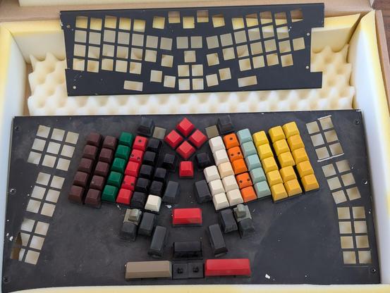 Picture of an ergonomic keyboard with unmarked keys of various colors. There are two thumb levers