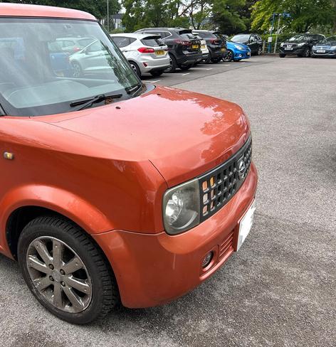 Front of an orange Nissan Cube Z11 in a car park, with the drivers window open and the hazard lights on, being loaded up with fursuits and luggage.