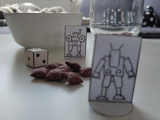 Two robot standees looking at each other high noon style, there's a cut and glued paper die, scattered chocolate cereal and a white cereal bowl.