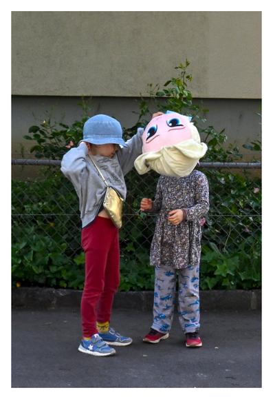 The image depicts two children outdoors, standing near a fence with greenery in the background. The child on the left is wearing a light gray long-sleeve shirt, red pants, blue shoes, and a blue bucket hat. They are also carrying a small gold crossbody bag. The child on the right is dressed in a dark floral-patterned dress over gray pants with blue patterns, and red shoes. The child on the left holds a large puppet head with blonde hair, covering the head of the other child. (created with help of Mistral.ai)