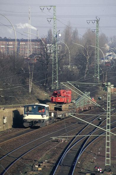 Eine kleine, altgediente Diesellok der Baureihe 332 (Köf III) in blau-beiger Bundesbahn-Lackierung zieht im gemächlichen Tempo einen kurzen Bauzug über ein mehrgleisiges Streckennetz. Der Zug besteht aus Flachwagen mit einem schweren, leuchtend roten Schienenkran und mehreren abgelegten Gleisjochen. Die Lok fährt auf einem Nebengleis, das leicht abschüssig aus einem kurzen Einschnitt herausführt. Der Lokführer ist gut erkennbar in seiner Kabine.

Im Vordergrund verzweigen sich die Hauptgleise mit mehreren Weichen und Signalen. Die Szene ist geprägt vom Gewirr der Oberleitungen und zahlreichen grün lackierten Gittermasten. Der Hintergrund zeigt eine dichte städtische Bebauung mit Industriearchitektur, Fabrikschornstein und Häusern hinter kahlen Winterbäumen. Im Dunst des Vormittags steigt Rauch auf – es herrscht typische Winterruhe auf einer norddeutschen Hauptbahnstrecke.
