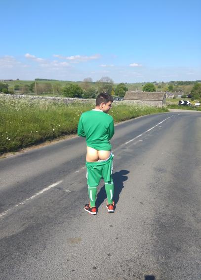 A person (it/its) stood in the middle of a road in the countryside, wearing a green football kit, with shorts shorts pulled down to reveal its butt.