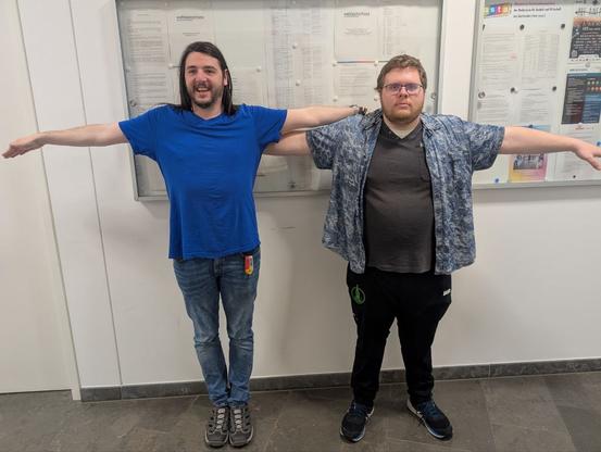 Zwei Dullies stehen in T-Pose vor einer Glasvitrine, in der die neue Wahlordnung aushängt
