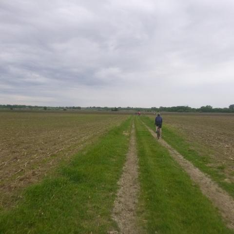 A wide angle view of a gravel farm path between two newly planted fields.  On either side of the path a small swath of green grass lines the tire tracks.  In the middle distance a single bicycle with a rider in blue is visible.  In the far distance a group of riders can just be seen as small dots.  The sky overhead is cloudy with the base of the clouds swirling and unsettled as rain showers were passing.