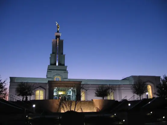 The image depicts a temple, specifically a building that is characteristic of Latter-day Saint (Mormon) architecture. It features a prominent spire topped with a statue, likely an angel, which is common in such temples. The setting appears to be during twilight, with the sky transitioning to night. The temple is well-lit, highlighting its architectural details and entrance. The surrounding area includes trees and steps leading up to the entrance.

Image Credits: Wikimedia / w:en:User:Rockford1963 / Public domain