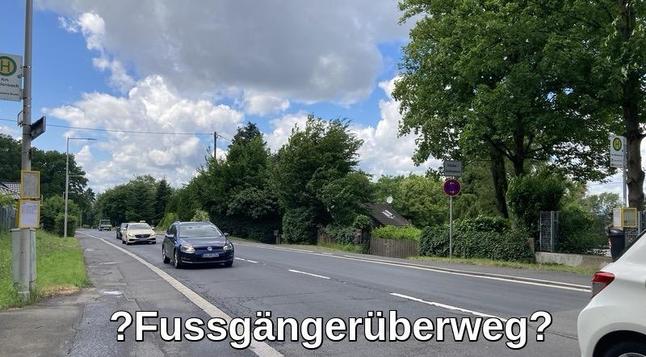 Rechts eine Bushaltestelle, dann folgt der Randstreifen der zweispurigen Straße, dann ein Bürgersteig, dann wieder eine Bushaltestelle. Ein schwarzes Auto kommt von unten, ein heller PKW hinterher. Links unten im Bild noch die Ecke eines weißen PKW zu sehen. Der Himmel ist bewegt bewölkt mit blauen Feldern. Rechts ein Baum. Im Hintergrund sind auch noch mehr Bäume zu sehen.