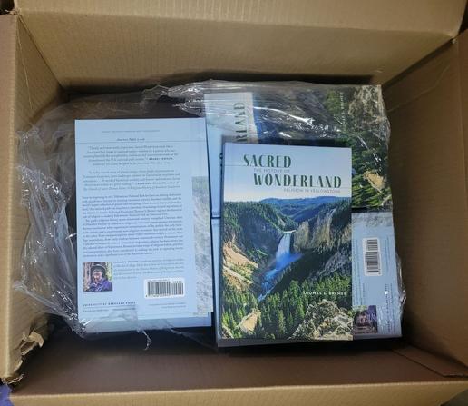 Open cardboard box containing multiple copies of the book Sacred Wonderland: The History of Religion in Yellowstone by Thomas S. Bremer. The book cover shows a photo of Yellowstone’s Lower Falls and canyon.