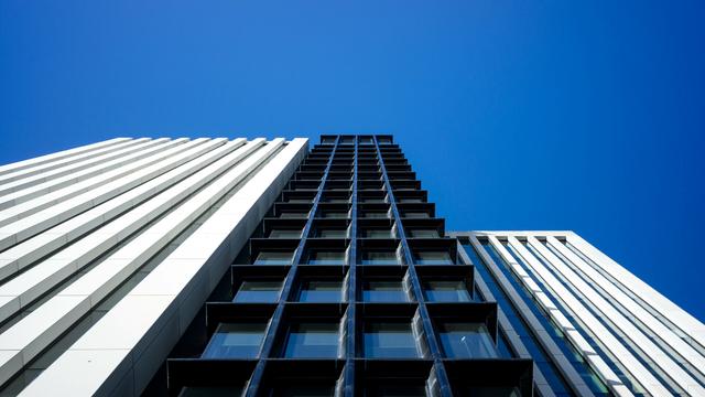 Photo looking up from near the base of a tall black building between two tall white buildings. The black building is a narrow four-wide grid of deeply indented windows. Both white buildings have thin recessed columns of windows running their entire height, the one on the right is set back a little. the sky above is clear bright blue.
