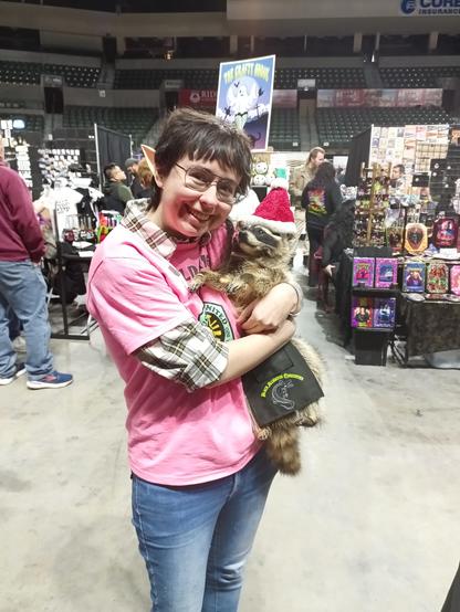 A girl with elf ears wearing a pink shirt and blue jeans is holding a taxidermied raccoon with a Santa hat and little apron on