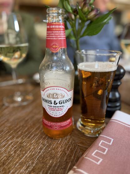 A clear glass bottle with a red neck label and a white front label. A Pilsner glass of light amber beer is to the right.