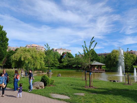 Parque con un estanque y unas fuentes a la derecha. Hay personas a la izquierda y árboles y edificios al fondo.