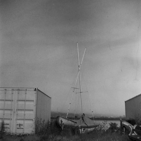 two small sailboats, on land, parked between shipping containers, with their masts crossed
