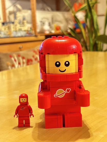 The image features two red LEGO astronaut figures: a large one made from LEGO bricks and a smaller traditional LEGO minifigure. Both have smiling faces and wear space-themed attire, with a planet logo on their chests.