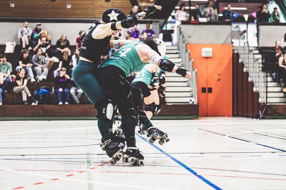 A color picture of two roller derby playing persons. On the left, in the air is a jamming person facing left like a bracket. On the right, on the ground, is a blocking person hitting the other person mid-air with their behind.