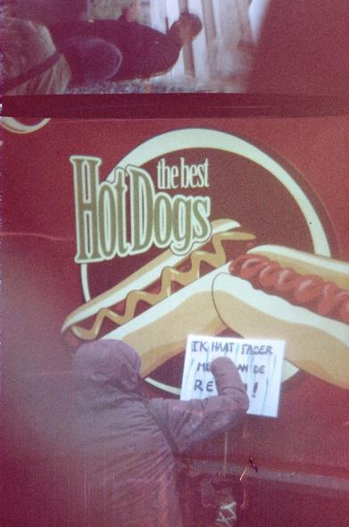 A woman in a rain coat paints a protest sign in Dutch, saying: "I hate Faber more than the rain". The sign leans against a hotdog stand that advertises with "The best hotdogs".